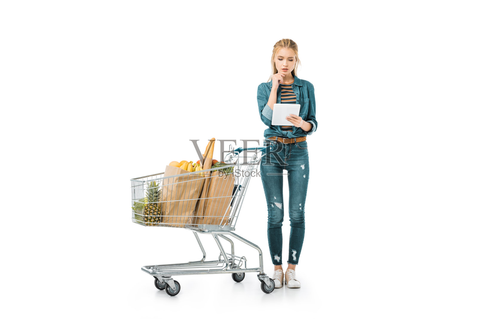 深思熟虑的女人看着数字平板电脑和站在购物推车与产品隔离在白色照片摄影图片