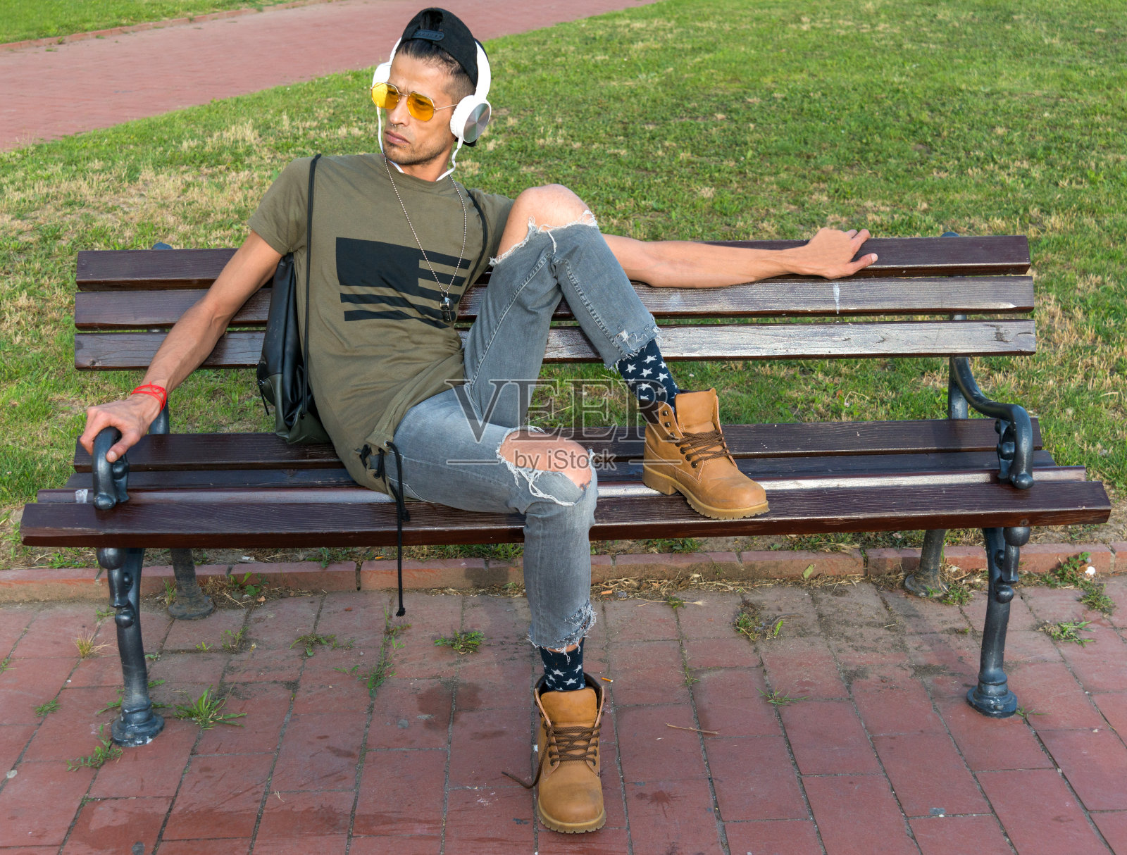 奢侈的(时髦的)男模特戴着太阳镜，耳机和黑帽子。一个男人坐在木凳上听音乐照片摄影图片