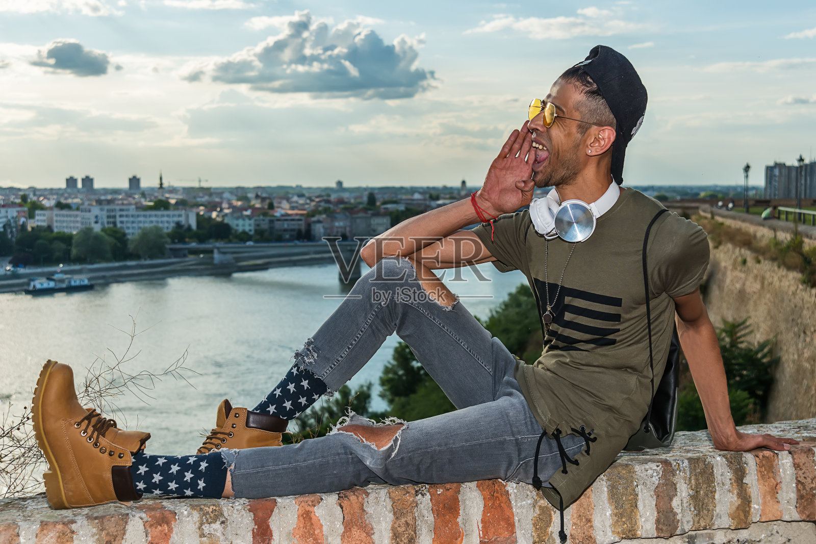 奢侈的(时髦的)男模特戴着太阳镜，耳机和黑帽子。背景是多瑙河和诺维萨德市。照片摄影图片