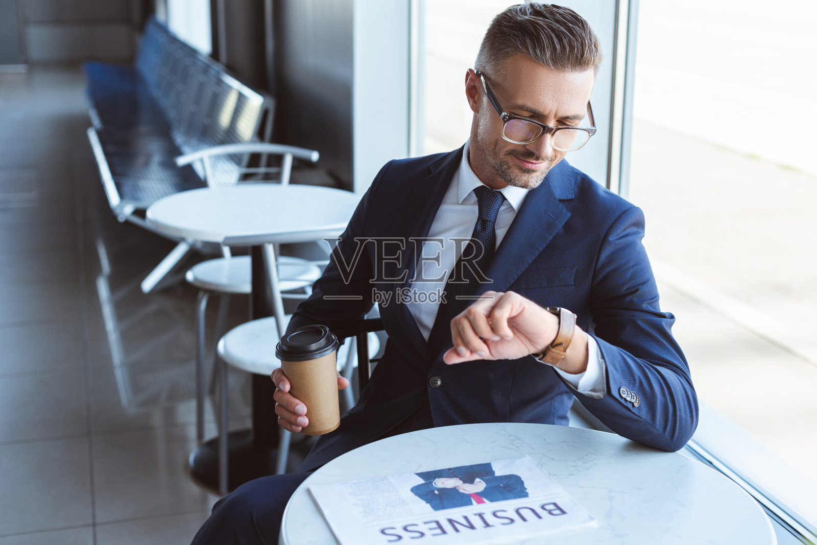 英俊的商人戴着眼镜拿着咖啡走，看着手上的手表照片摄影图片