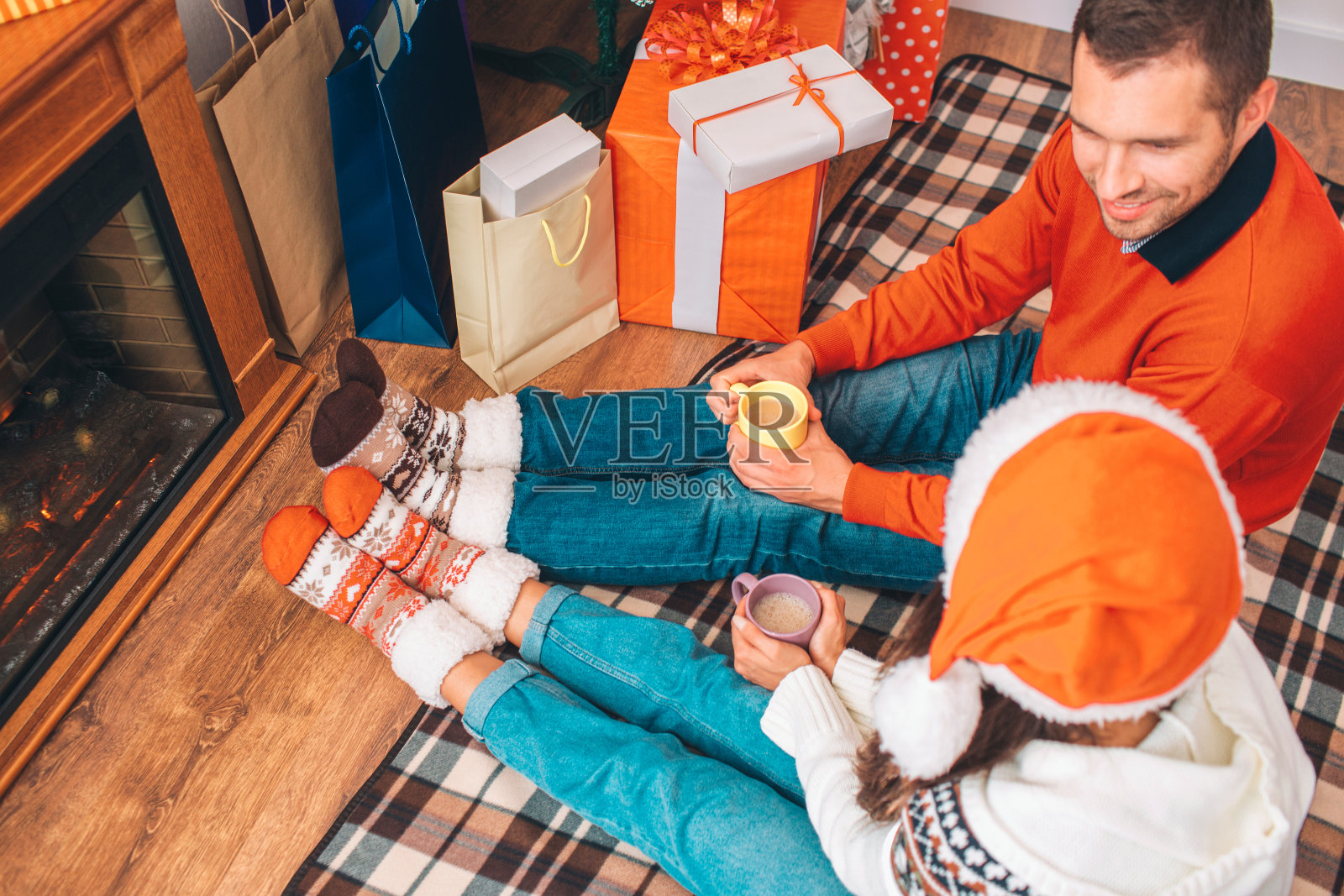 一对夫妇一起坐在壁炉边的地板上。他们拿着几杯可可。人们互相看着，微笑着。他们是快乐的。照片摄影图片