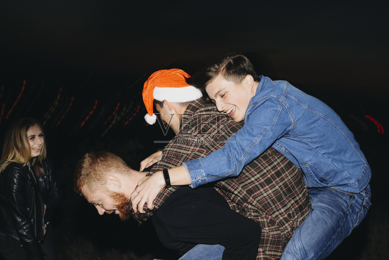 一个兴奋的年轻人背着两个男性朋友在一个女人附近庆祝圣诞节的夜晚。背着朋友的人照片摄影图片