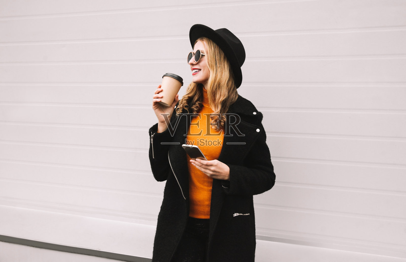休息时间!在灰色的墙壁背景下，一个微笑着的时尚女人拿着智能手机在城市里喝着咖啡照片摄影图片