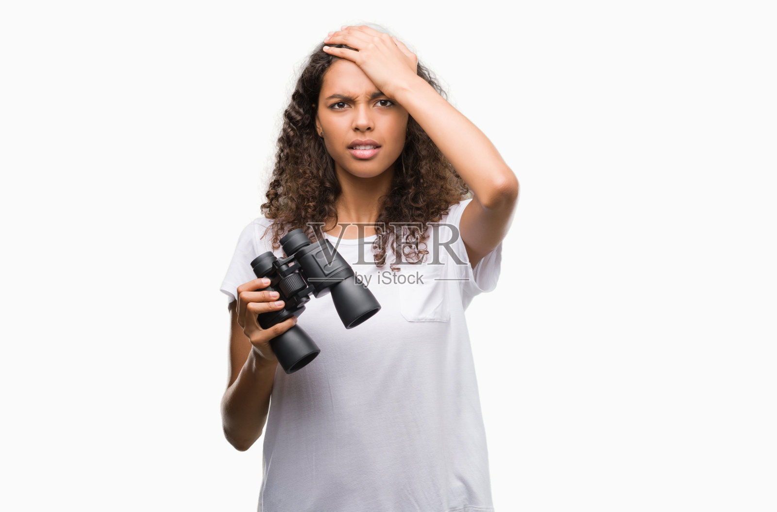 年轻的西班牙妇女通过双筒望远镜看着强调与手在头上，震惊与羞愧和惊讶的脸，愤怒和沮丧。为错误而恐惧和不安。照片摄影图片