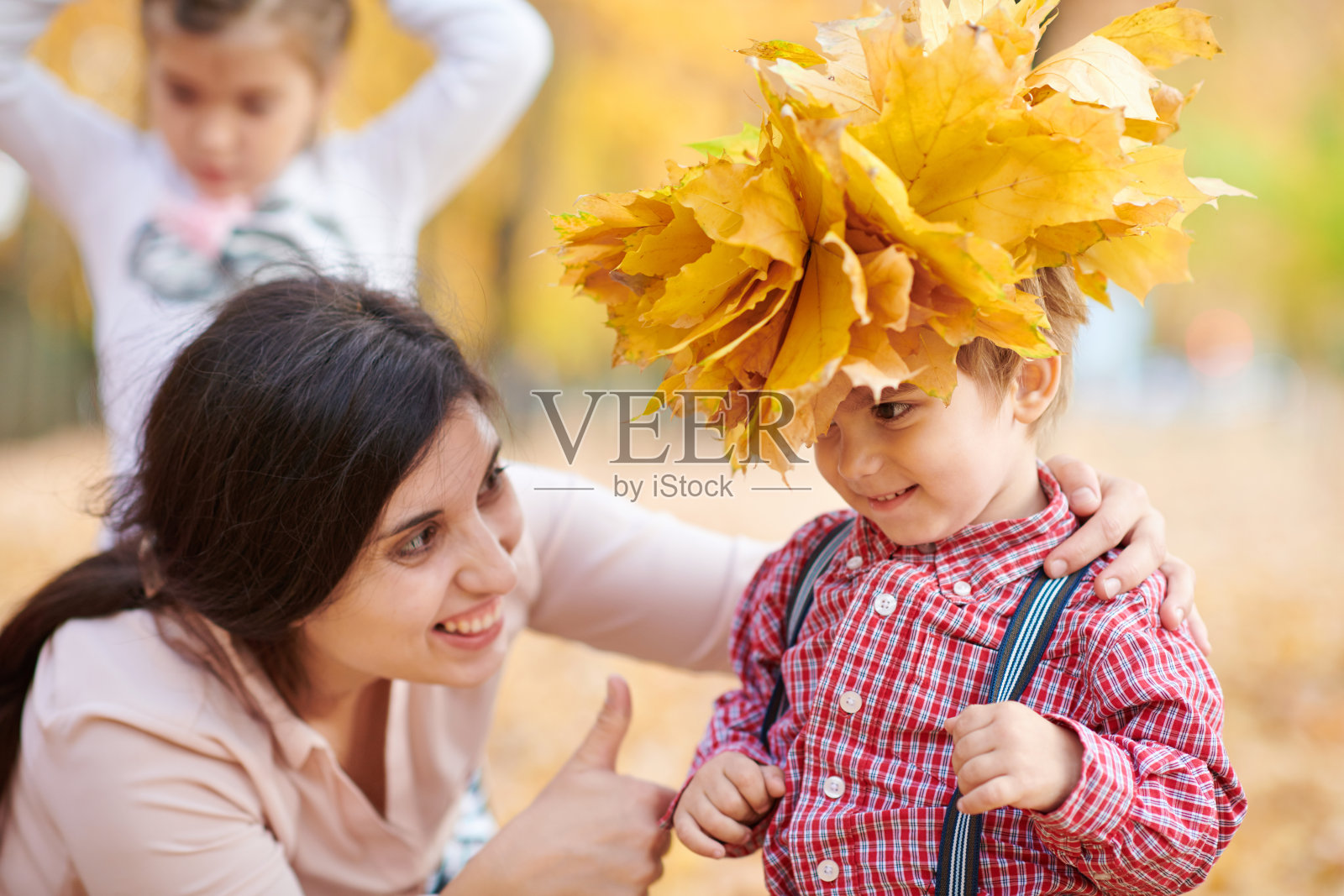 妈妈把黄色的落叶戴在儿子头上。幸福家庭在秋城公园。孩子和父母。他们摆姿势，微笑，玩耍，玩得很开心。亮黄色的树木。照片摄影图片