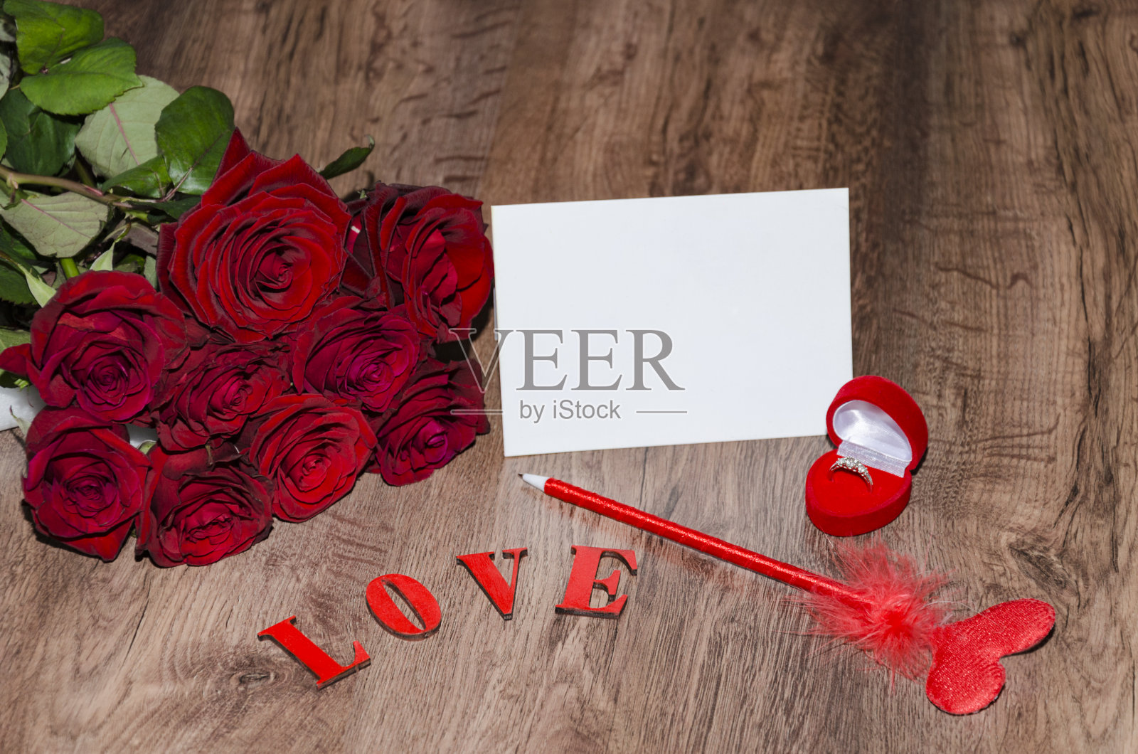 棕色的木桌，一张白色的明信片，一个提手，盒子里有一个戒指，上面装饰着红色的玫瑰花，照片摄影图片