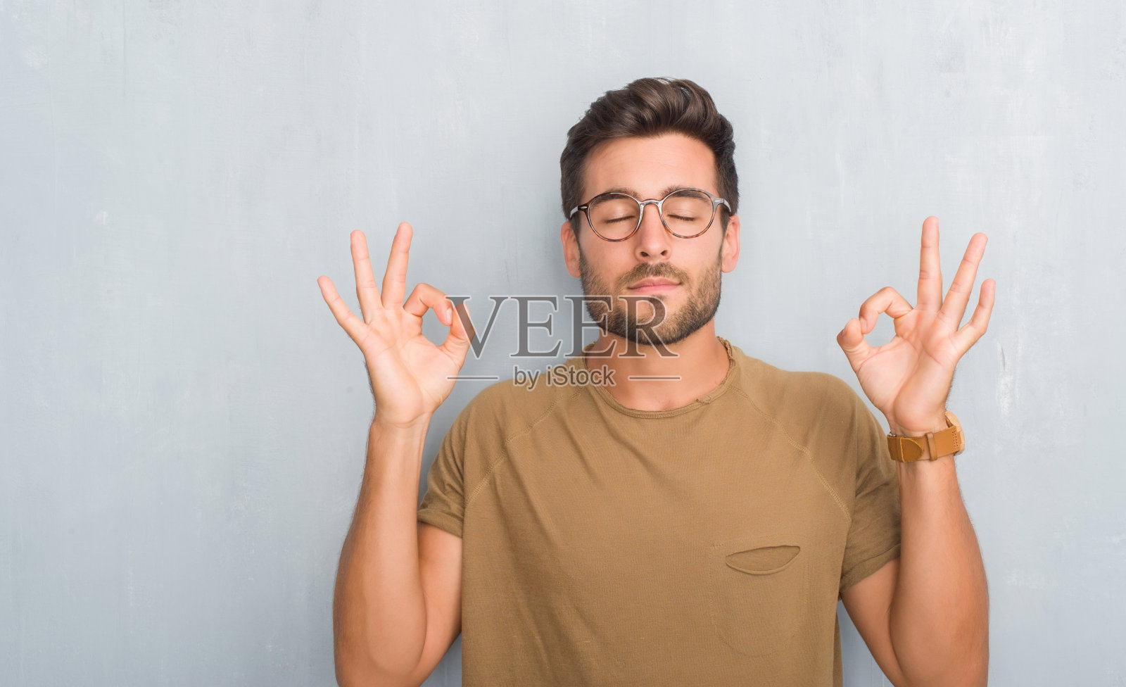 英俊的年轻人越过灰色垃圾墙戴眼镜放松和微笑闭上眼睛做冥想手势与手指。瑜伽的概念。照片摄影图片