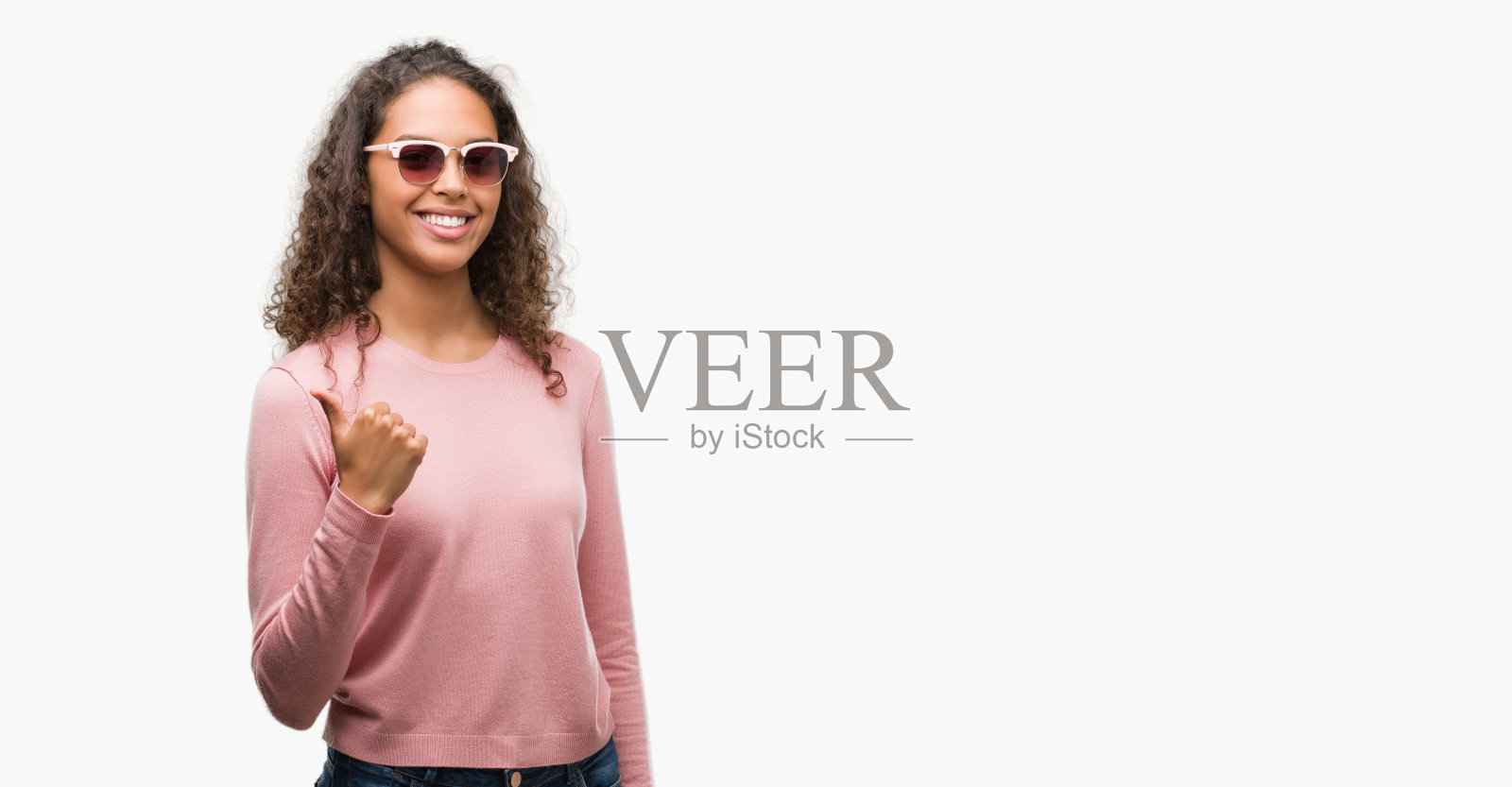 美丽的年轻西班牙妇女戴着太阳镜做快乐的拇指手势与手。赞许的表情看着镜头，表示成功。照片摄影图片