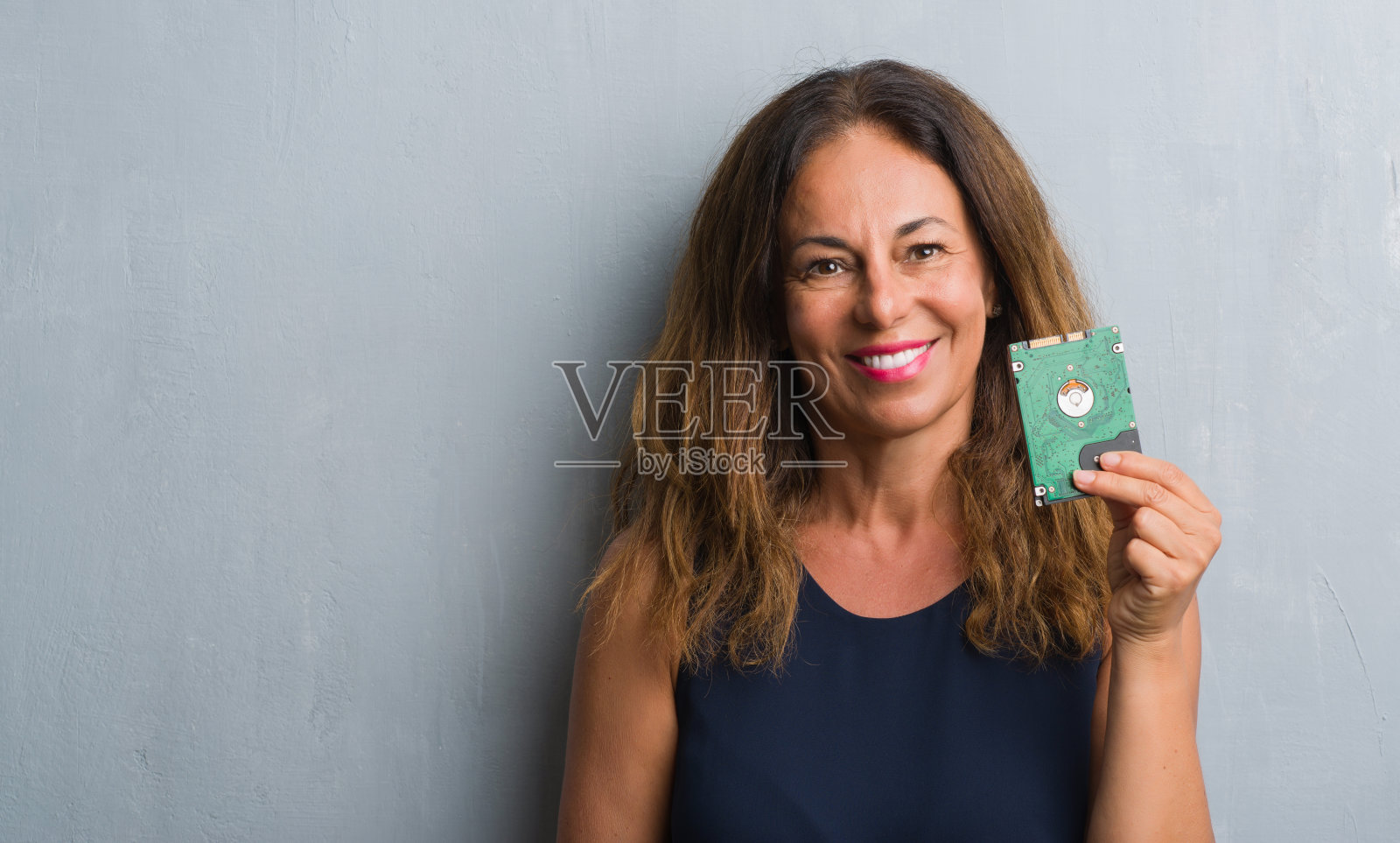 中年拉美裔妇女站在灰色垃圾墙持有硬盘与一个快乐的脸站和微笑与自信的微笑显示牙齿照片摄影图片