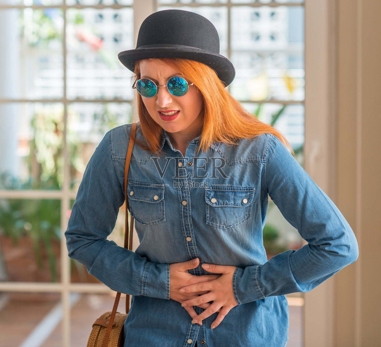 时髦的红发女人戴着圆顶礼帽和太阳镜，手放在肚子上，因为消化不良，痛苦的疾病感觉不舒服。疼痛的概念。照片摄影图片
