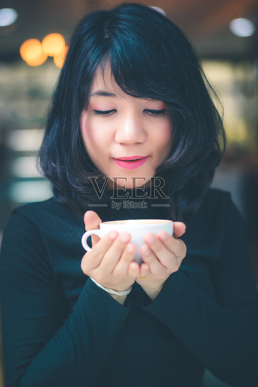 美丽迷人的年轻亚洲女子捧着一杯咖啡或手里拿着咖啡坐在咖啡馆的沙发上，复古的色调。照片摄影图片