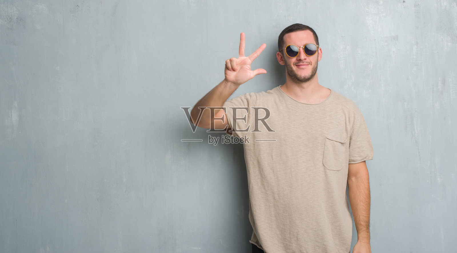 年轻的白人男子越过灰色的垃圾墙戴着太阳镜显示和指向第三个手指，同时自信和快乐的微笑。照片摄影图片