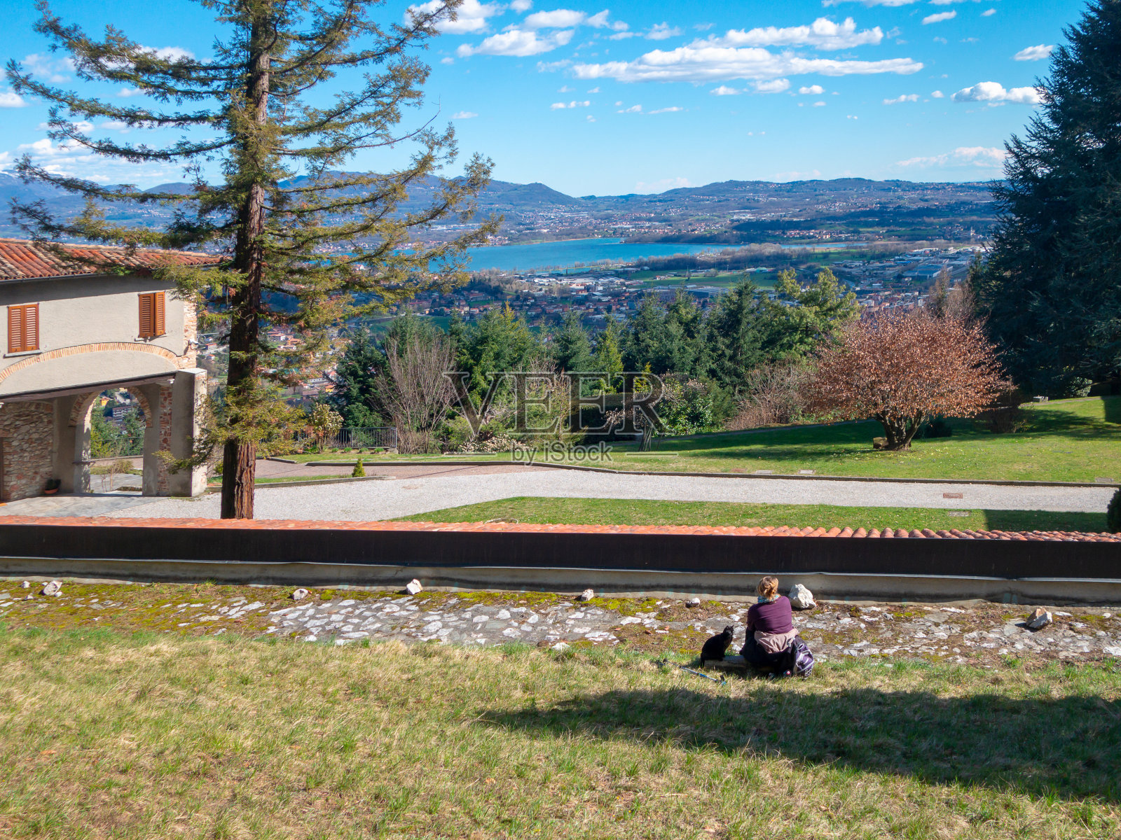 女人在一次爬山后休息，坐在公园的草地上的一个女修道院，俯视着阿尔卑斯山的湖、山、村的全景。照片摄影图片