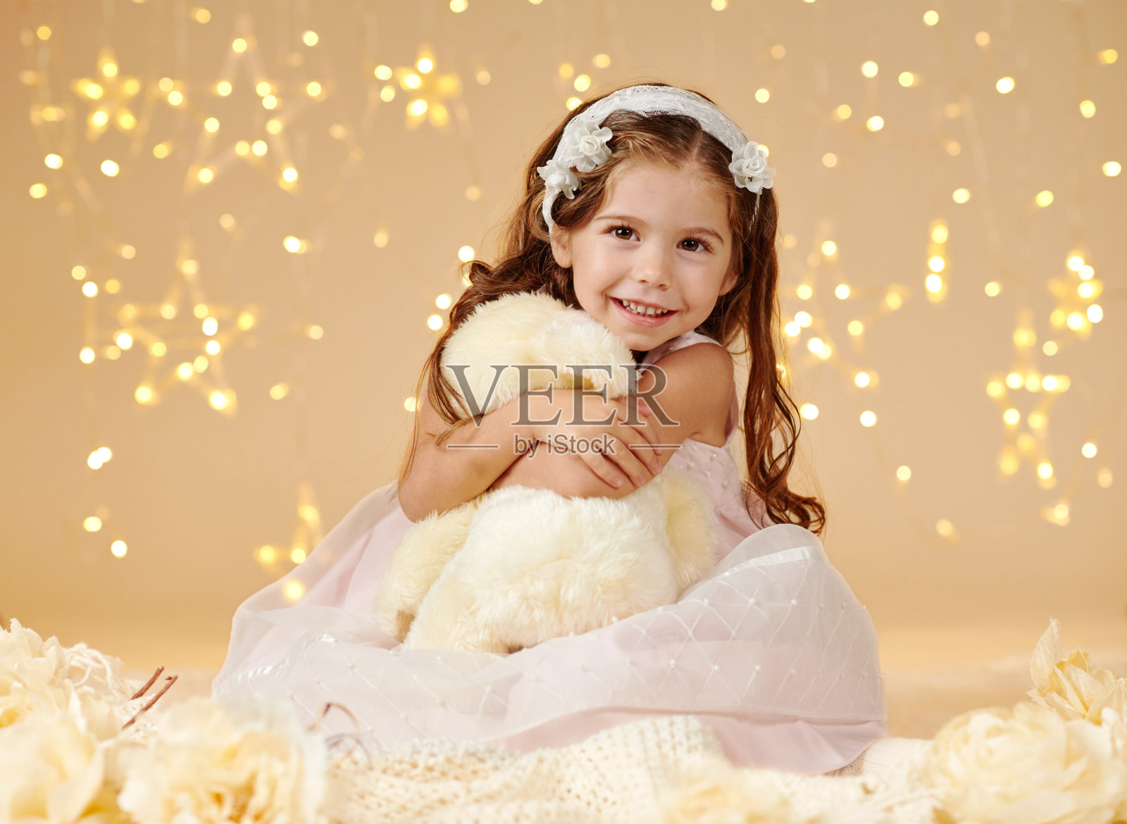 小女孩带着玩具熊在圣诞彩灯下摆姿势，黄色的背景，粉色的裙子照片摄影图片