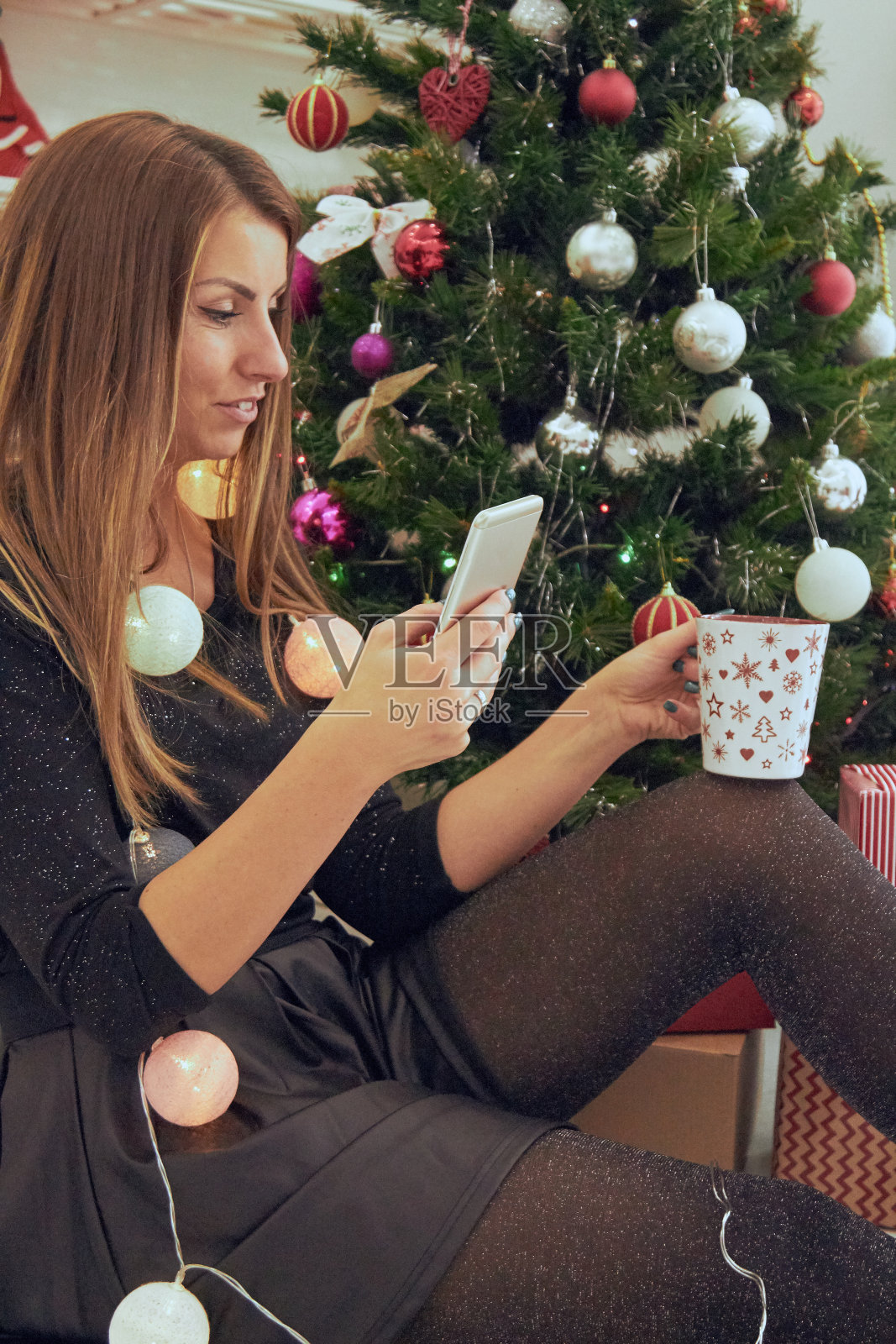 美丽的女人使用手机和喝咖啡接近圣诞夜/新年。照片摄影图片
