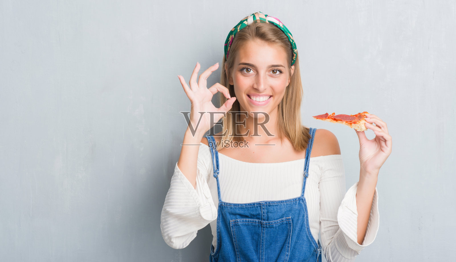 美丽的年轻女子在垃圾灰墙吃辣香肠披萨片做ok手势与手指，优秀的象征照片摄影图片