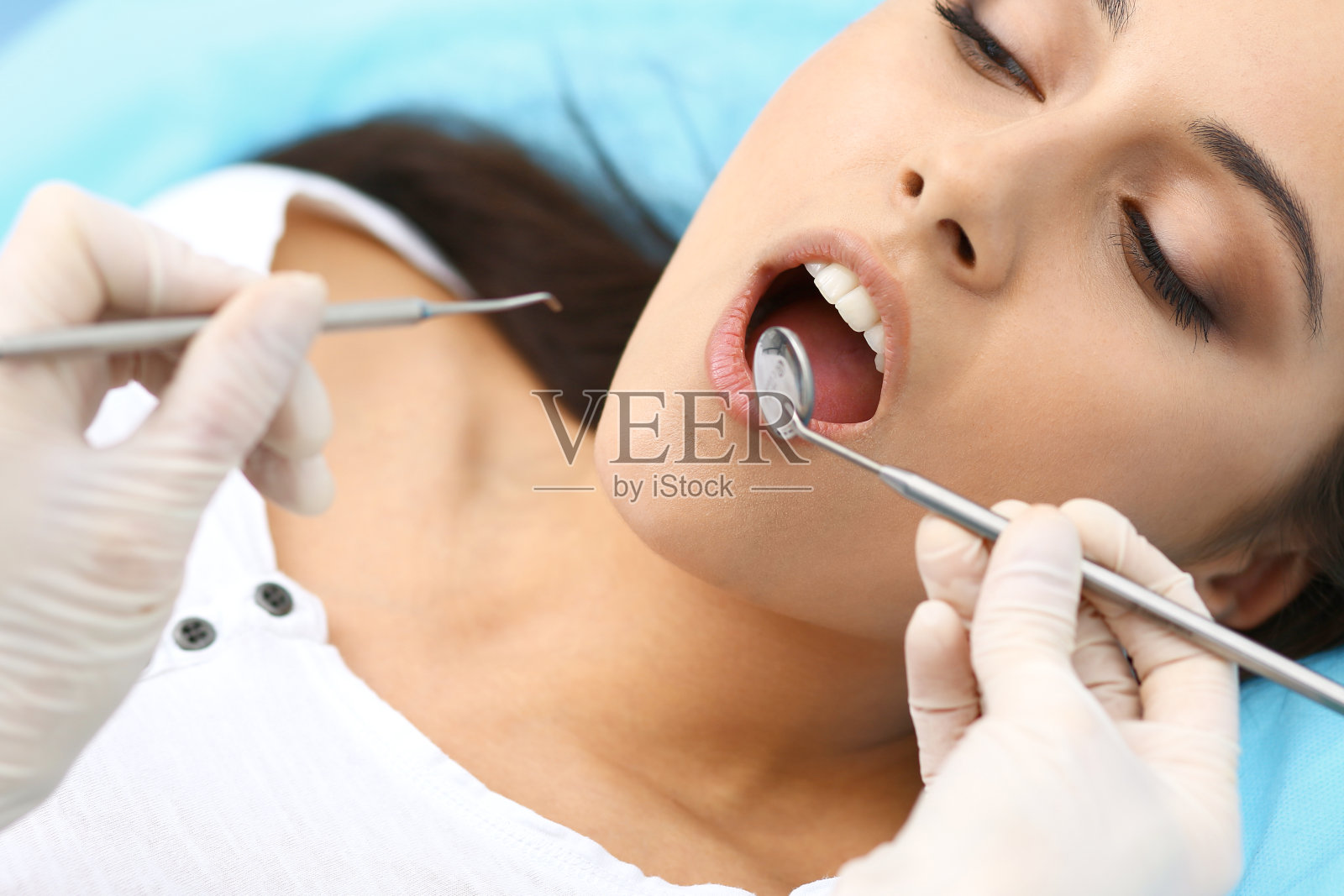 年轻的女病人正在看牙医。美丽的女人与健康的直白牙齿坐在牙科椅子与张大的嘴在口腔检查期间，而医生工作的牙齿照片摄影图片