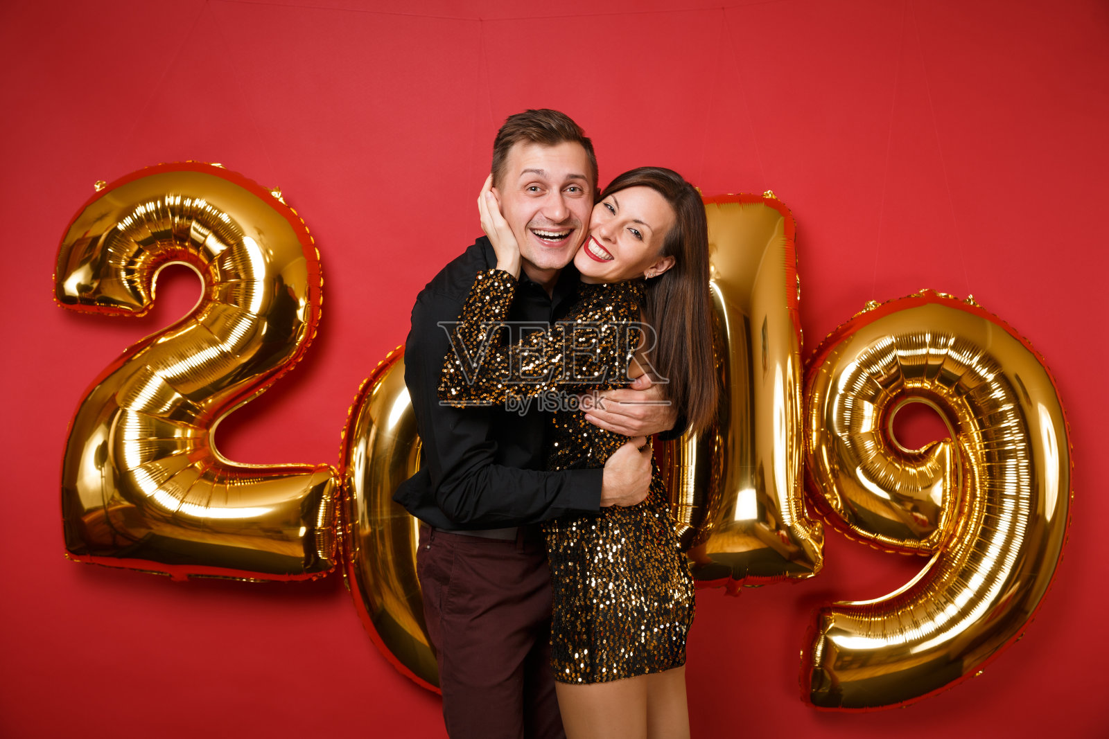 一对男孩女孩穿着闪闪发光的裙子，黑色衬衫庆祝节日派对孤立在明亮的红色墙壁背景金色数字气球工作室肖像。新年快乐2019圣诞概念。照片摄影图片