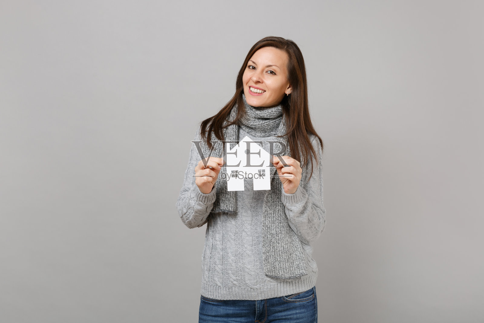 微笑的年轻女子在灰色毛衣，围巾拿纸白色的房子孤立在灰色的背景在工作室。健康时尚的生活方式，人们真诚的情感，寒冷的季节理念。模拟拷贝空间。照片摄影图片