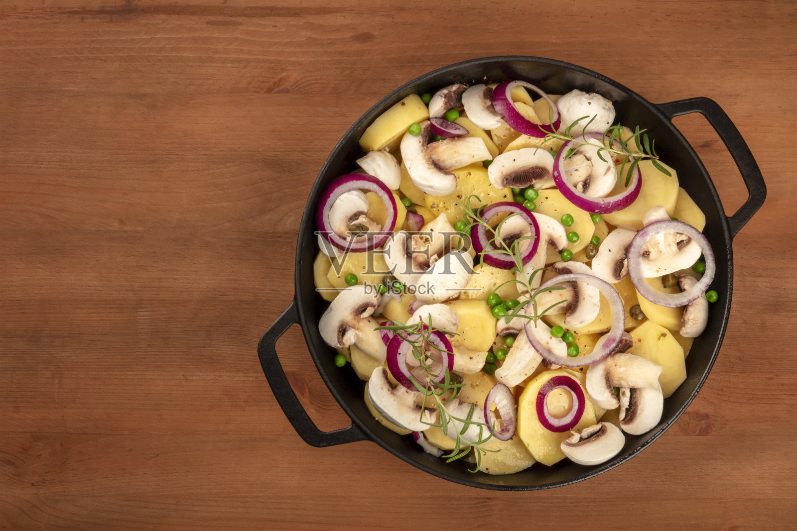一个装有生蔬菜、土豆、洋葱、绿豌豆、蘑菇和迷迭香的平底锅，放在一张黑色的乡村木桌上，上面放着文字照片摄影图片