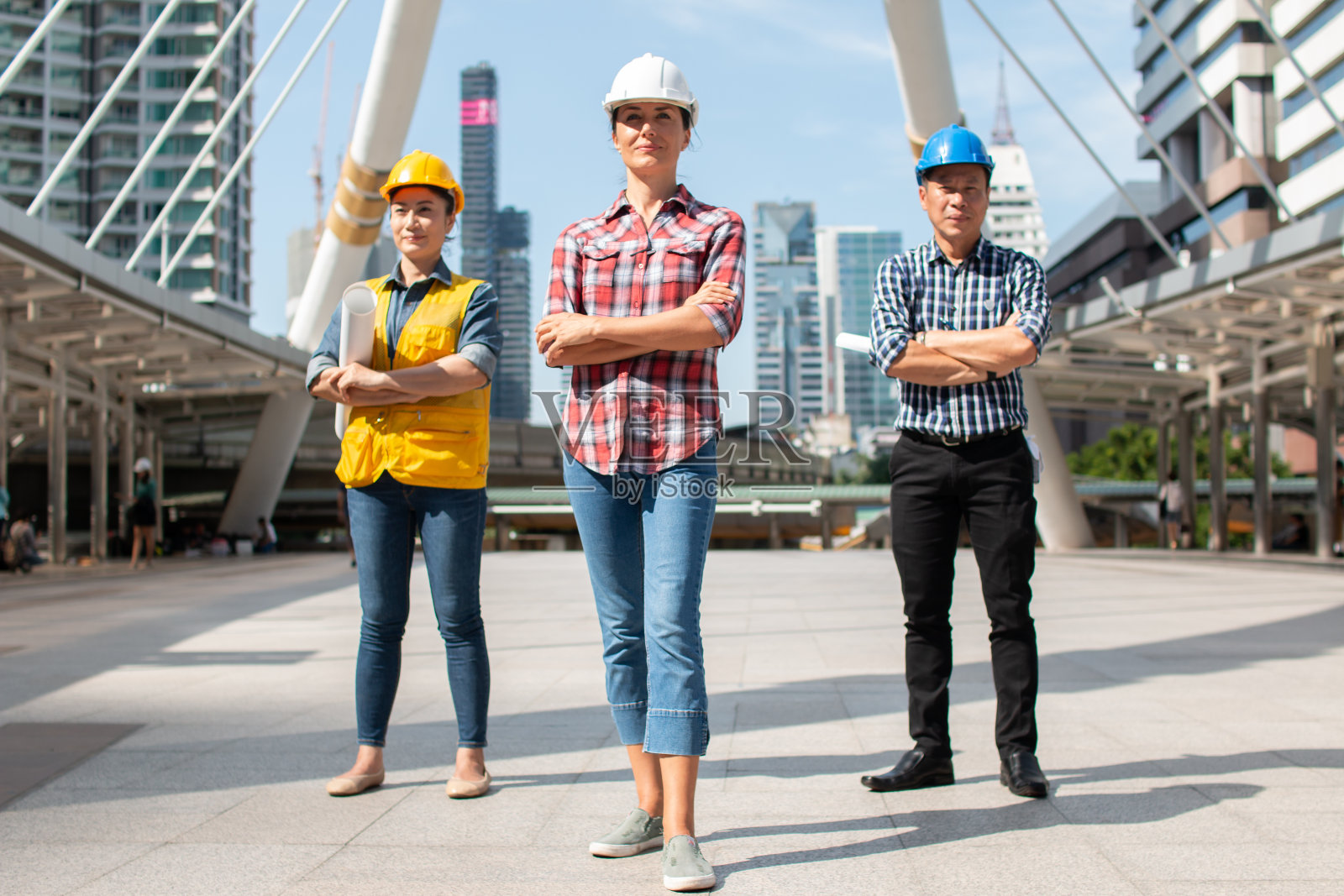 三名工业工程师戴着安全帽，双手交叉站在办公室外的大楼上，手拿检查牌，面带微笑。工程工具和施工概念。照片摄影图片