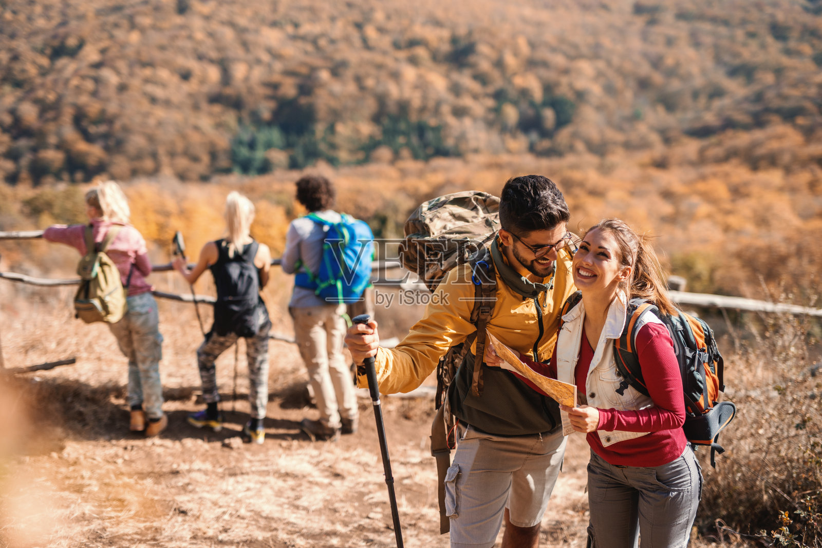 可爱的夫妇看着地图，而他们的朋友在背景看视图。徒步旅行的概念。照片摄影图片