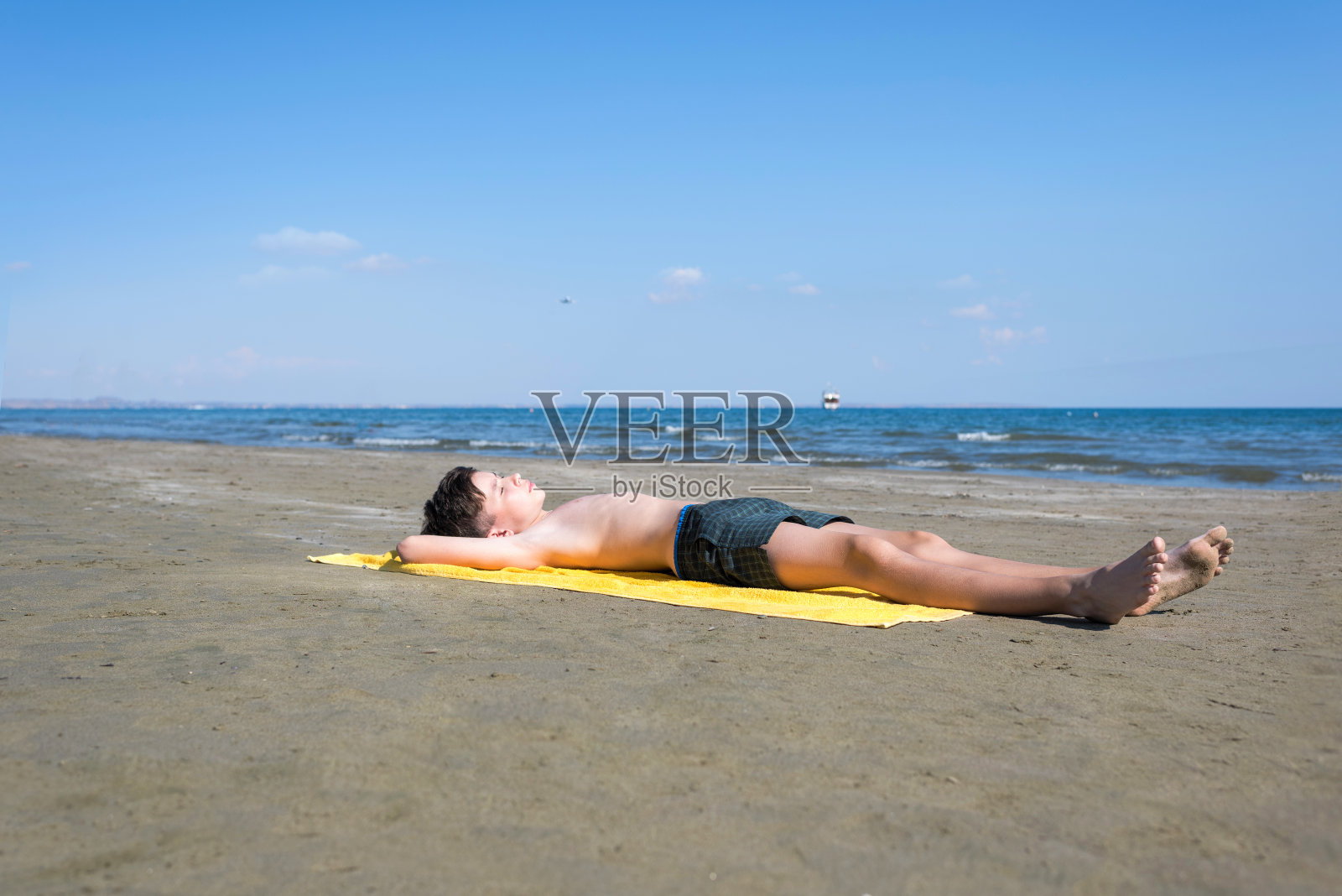 一个十几岁的男孩躺在沙滩上，躺在黄色的毛巾上晒太阳。和孩子们一起乘飞机旅行。Mackenzie海滩,拉纳卡、塞浦路斯照片摄影图片