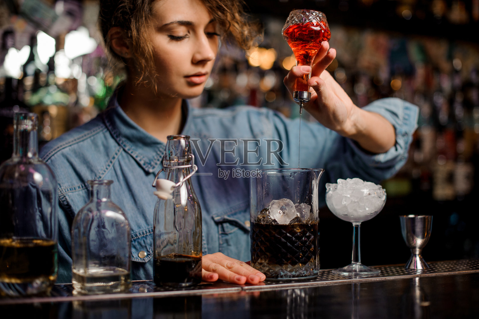 女调酒师从玻璃瓶中向装有冰块的量杯中倒入红色苦汁照片摄影图片