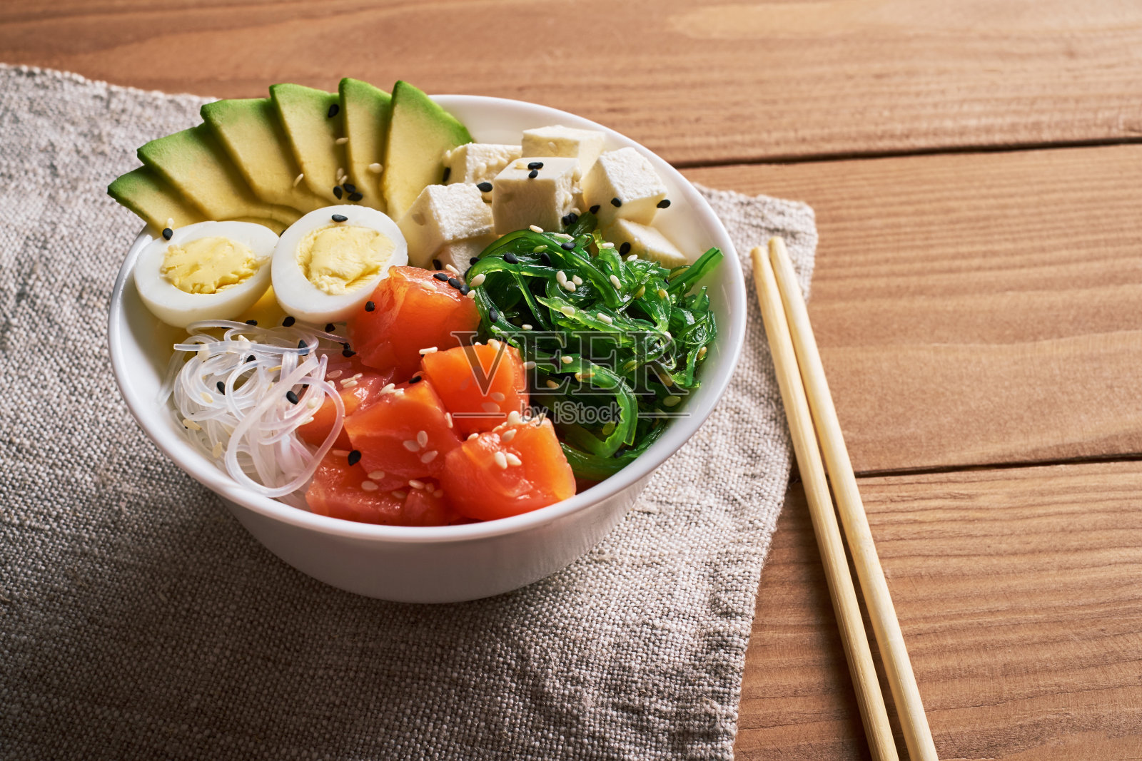 碗里有三文鱼、鳄梨、米饭、chuka沙拉、奶酪、鹌鹑蛋，洒上白芝麻和黑芝麻，配以筷子和酱油，亚麻餐巾桌布放在木质背景上照片摄影图片