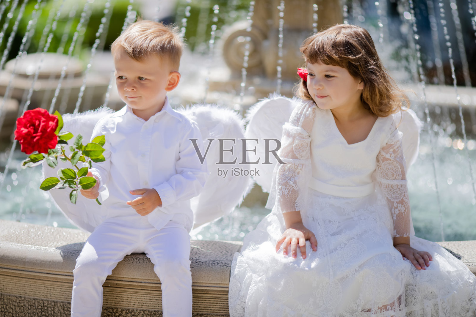 可爱的小男孩和小女孩穿着天使服装坐在一起，男孩拿着红玫瑰照片摄影图片