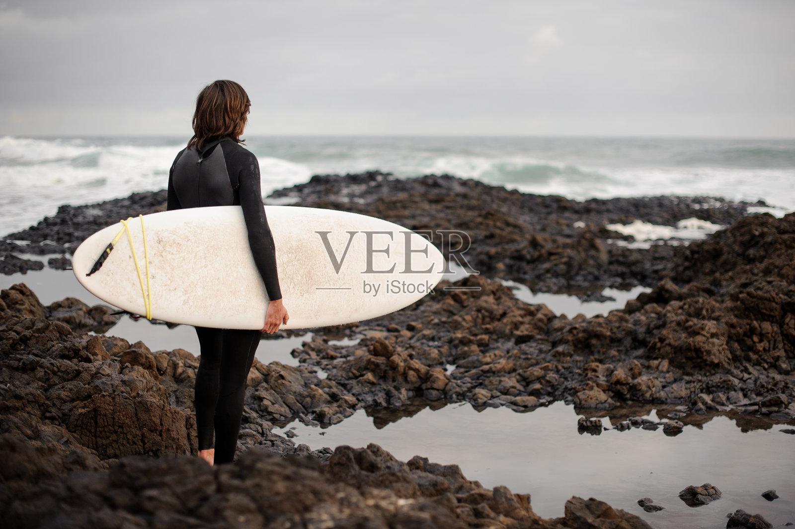 后视图黑发男孩站在黑色泳衣与冲浪在他的手在岸边照片摄影图片
