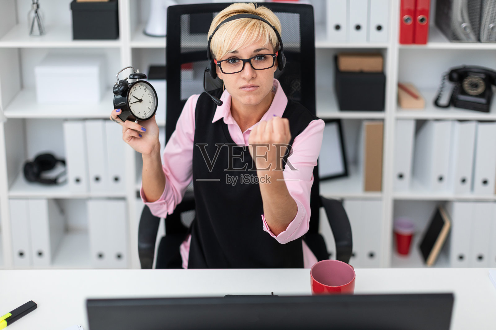 一个年轻的女孩坐在电脑桌前，手里拿着一个闹钟。照片摄影图片
