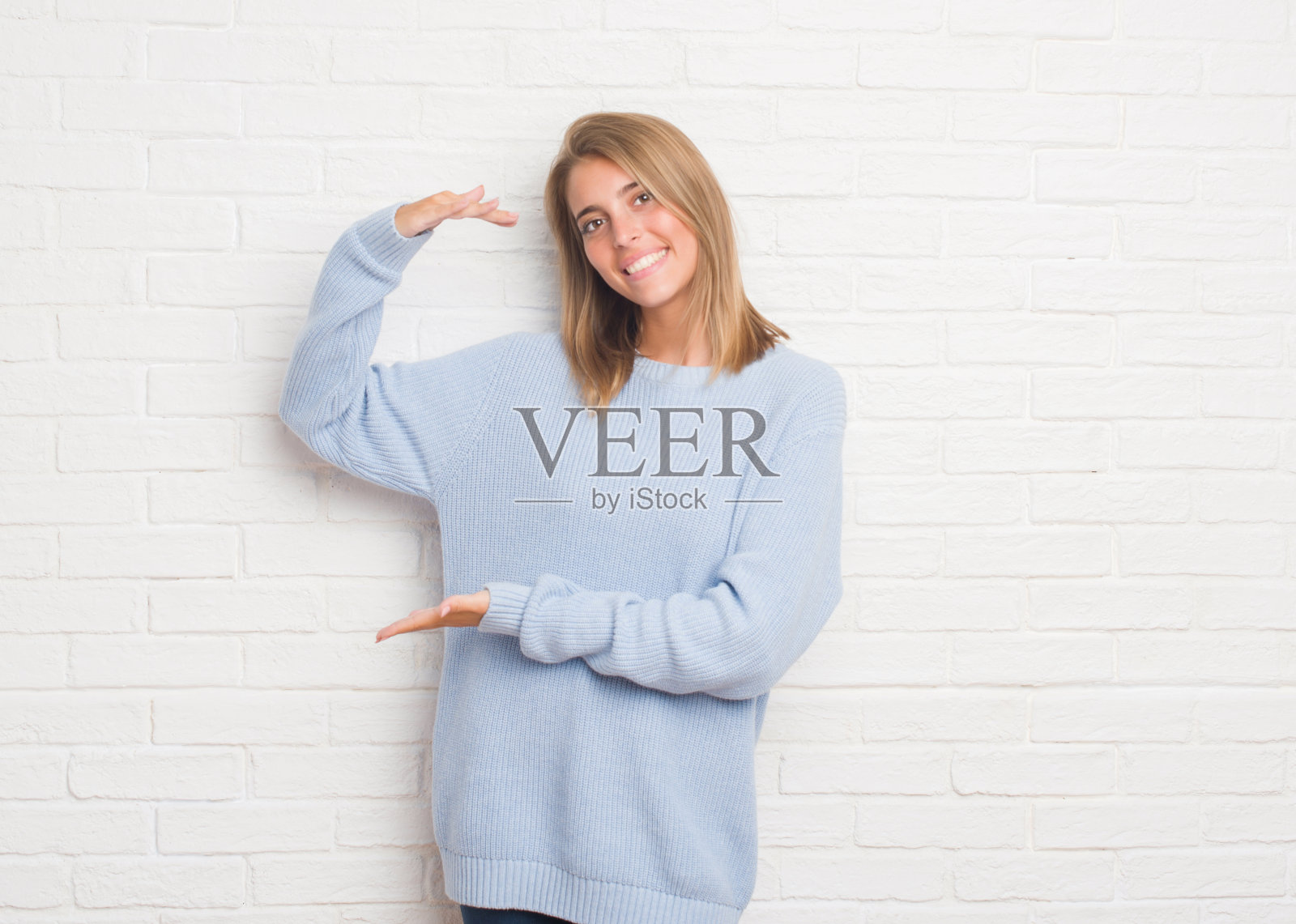 美丽的年轻女子在白砖墙上用手比划着大大的尺寸标志，测量符号。看着镜头微笑。测量的概念。照片摄影图片