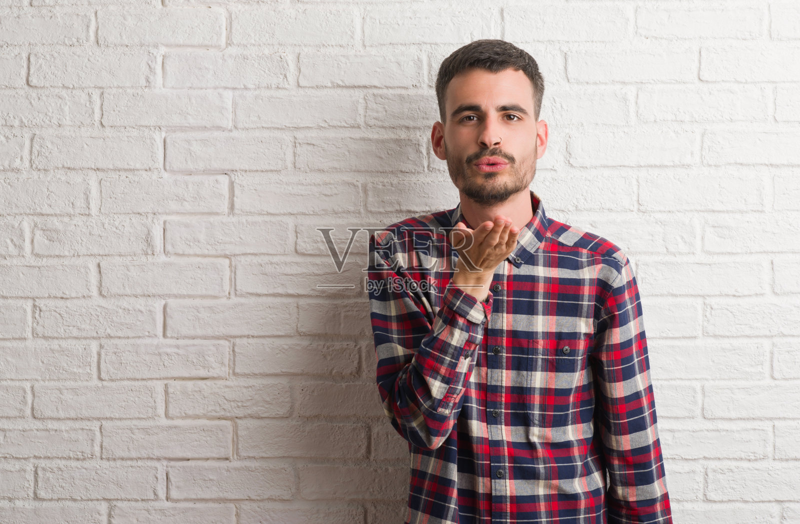 年轻的成年男子站在白色的砖墙看着镜头，用手在空中飞吻是可爱和性感的。爱的表达。照片摄影图片