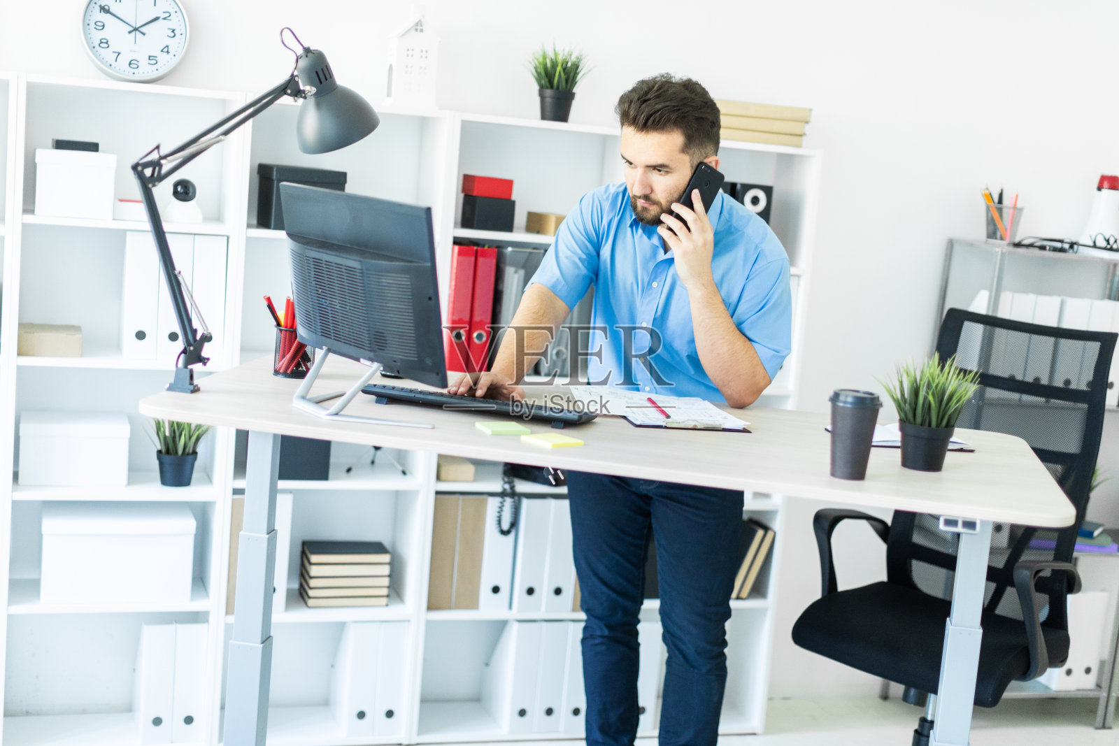 一个年轻人站在办公室的电脑桌前，正在讲电话。照片摄影图片