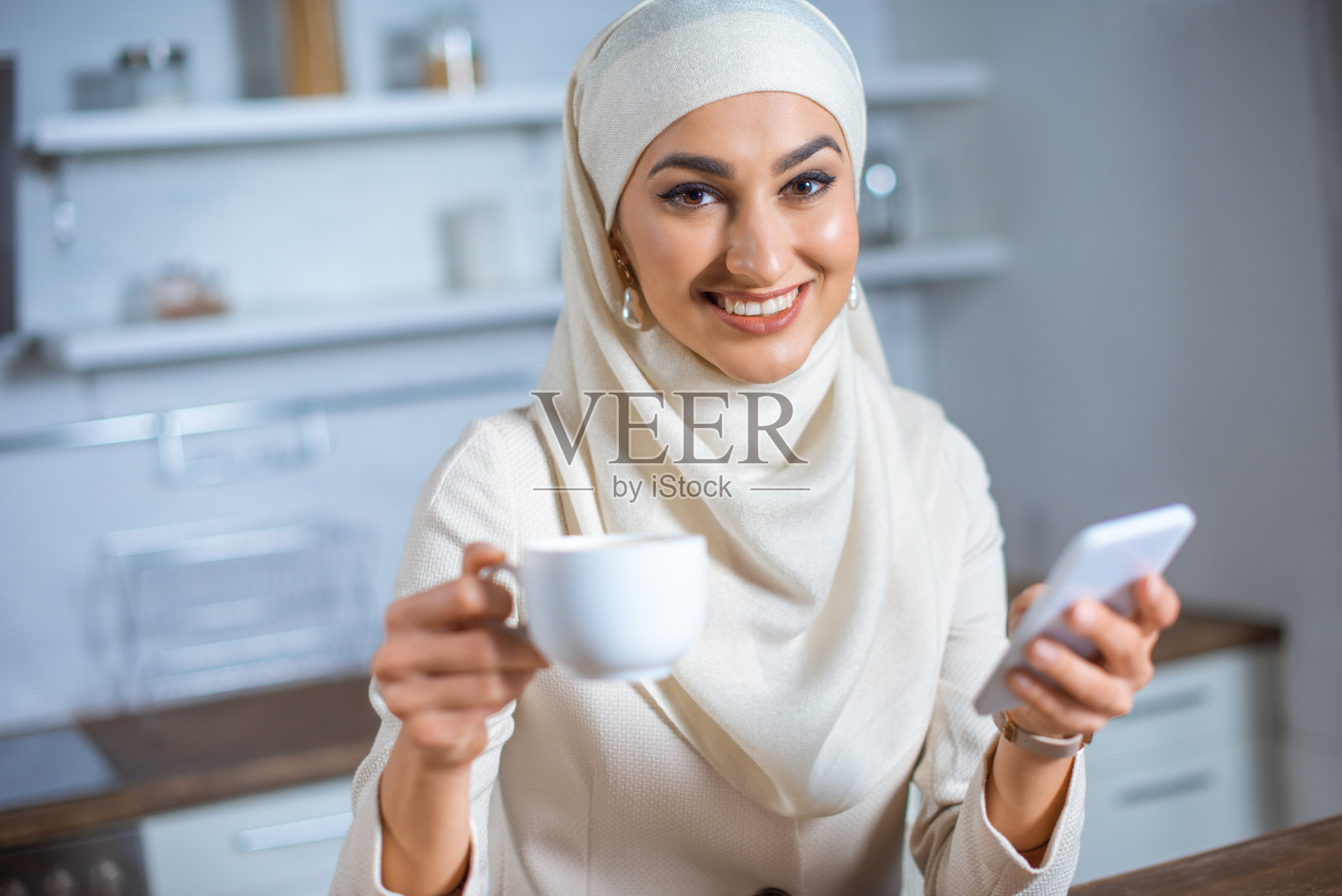 微笑的年轻穆斯林妇女拿着咖啡和智能手机在家照片摄影图片