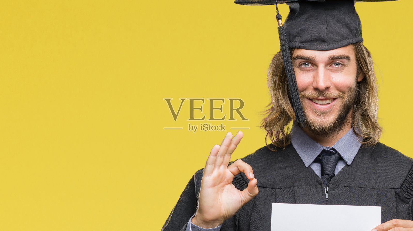 年轻英俊的毕业生与长头发拿着空白纸在孤立的背景做ok手势与手指，优秀的象征照片摄影图片