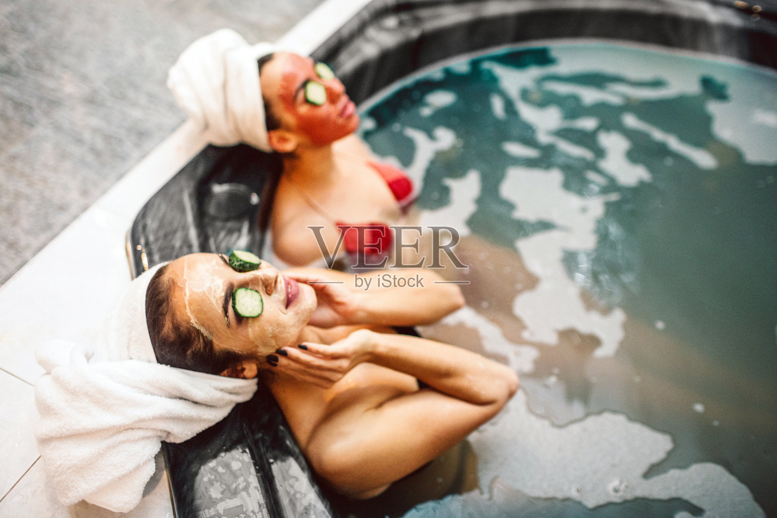 放松的年轻女性在热水浴缸里做水疗。它们的眼睛上方有面罩和几片黄瓜。模特们看起来既舒适又整齐。照片摄影图片