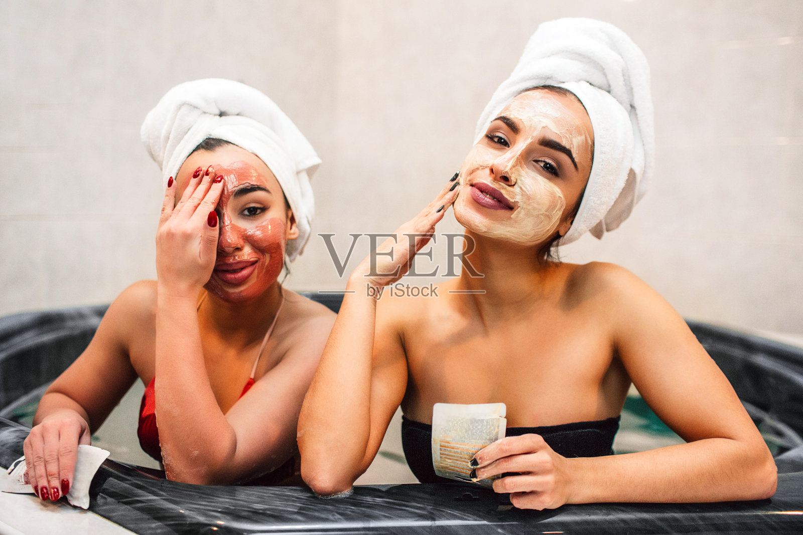 轻松的年轻女子看着镜头。他们的脸上戴着白色和红色的面具。模特们坐在装满水的热浴盆里。女人用泥面膜摸脸。照片摄影图片