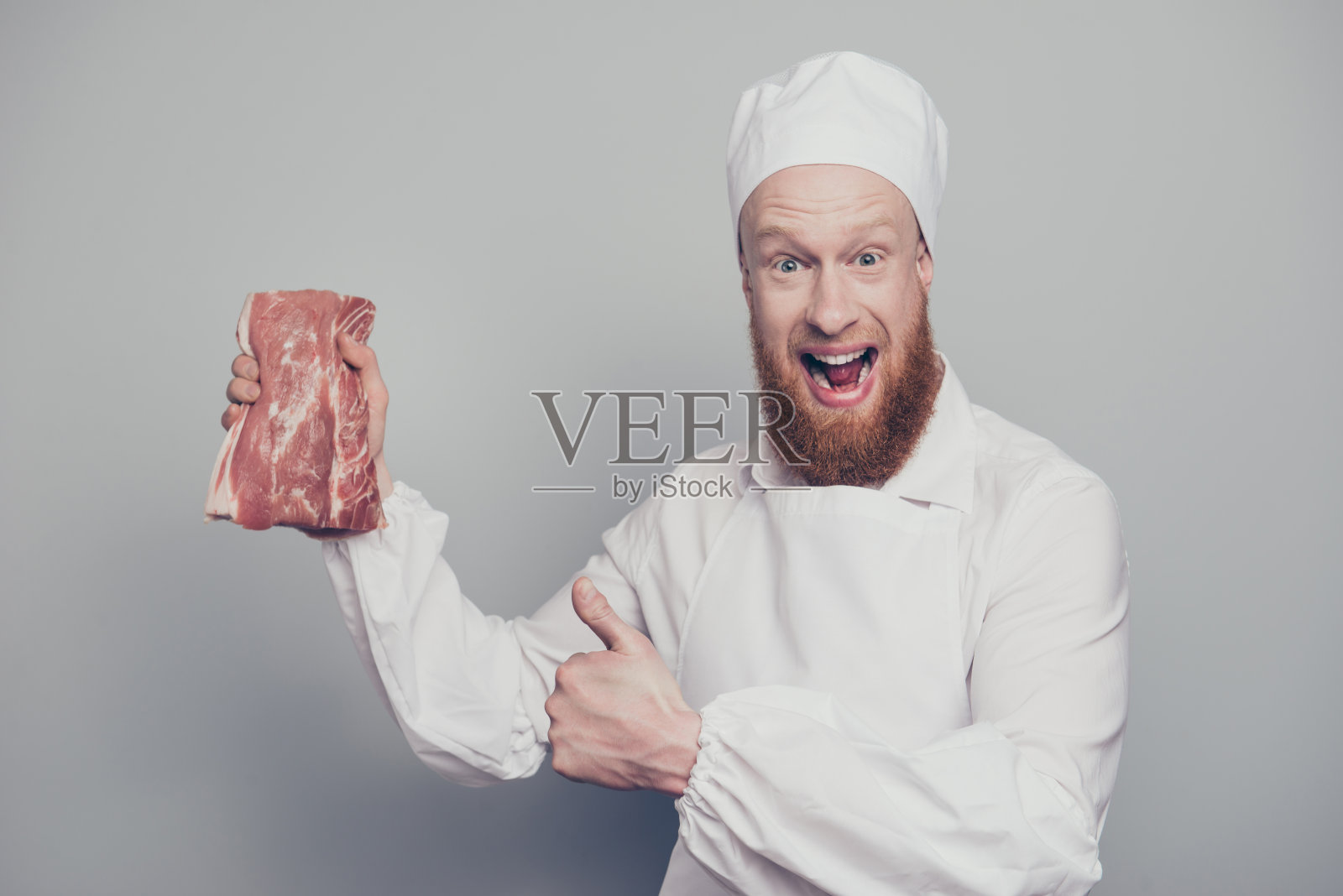 他的肖像，漂亮的疯狂有趣的屠夫与胡茬手握新鲜的肉在手臂大叫尖叫，张开的嘴显示大拇指孤立在灰色柔和的背景照片摄影图片