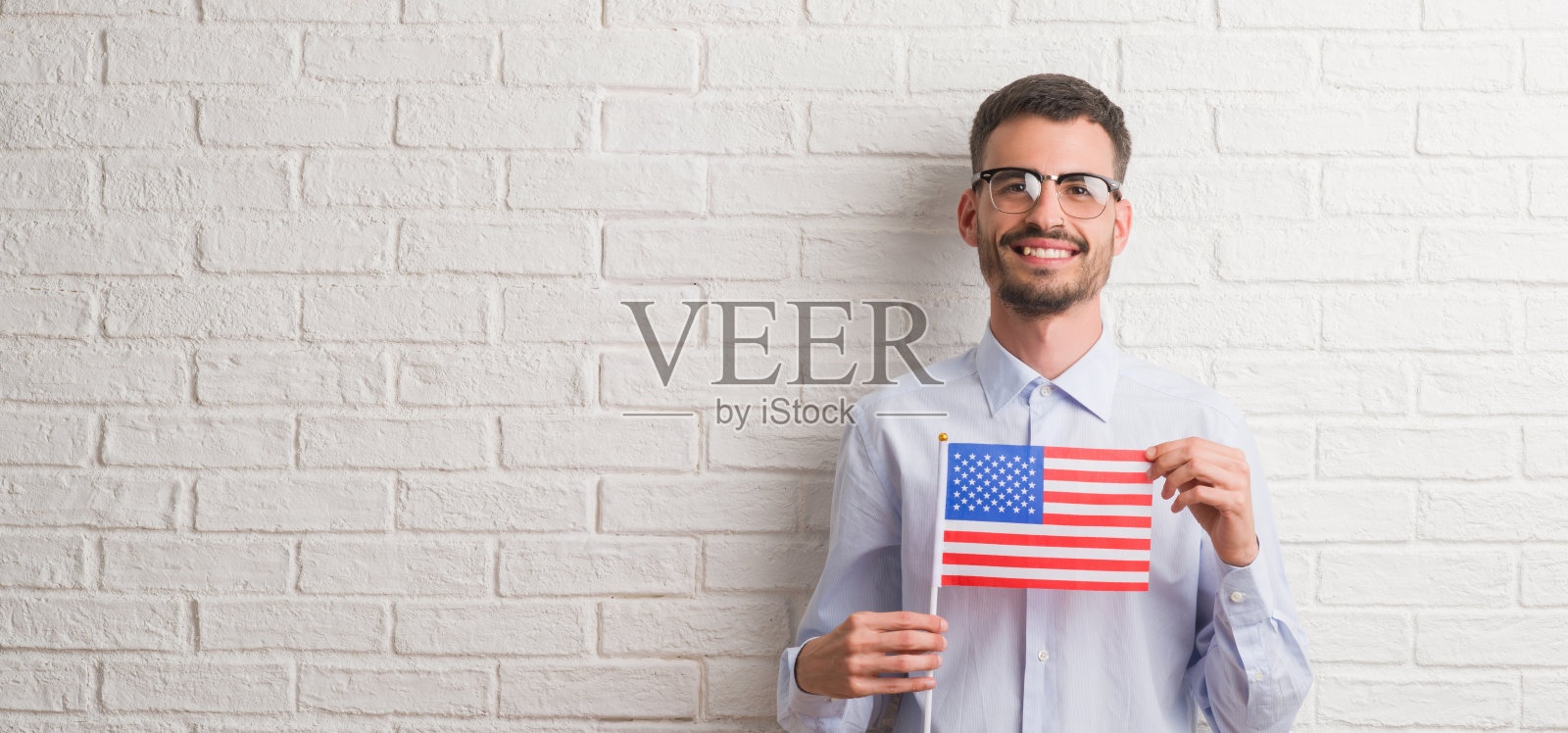 年轻的成年男子举着美国国旗越过砖墙，快乐地站着，自信地笑着露出牙齿照片摄影图片