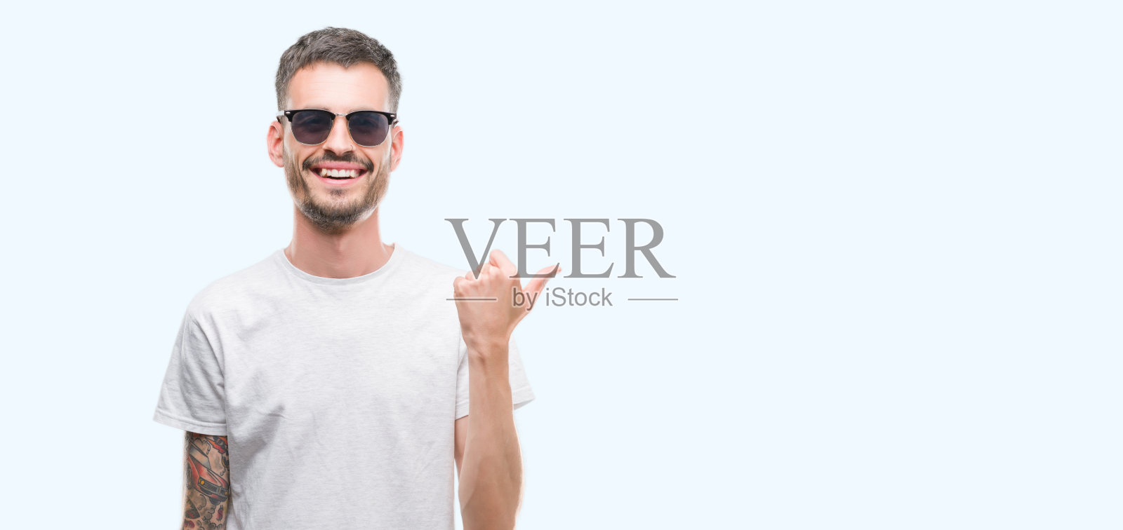 年轻的纹身成年男子戴着太阳镜指向和显示拇指向上的一边与幸福的脸微笑照片摄影图片
