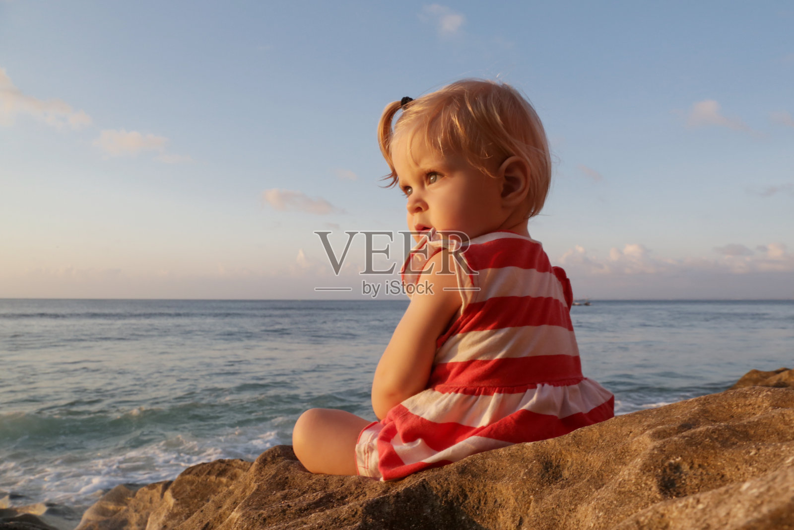 一岁的小女孩穿着红白相间的连衣裙坐在岩石海岸上，花时间在户外对小孩子来说是很重要的照片摄影图片