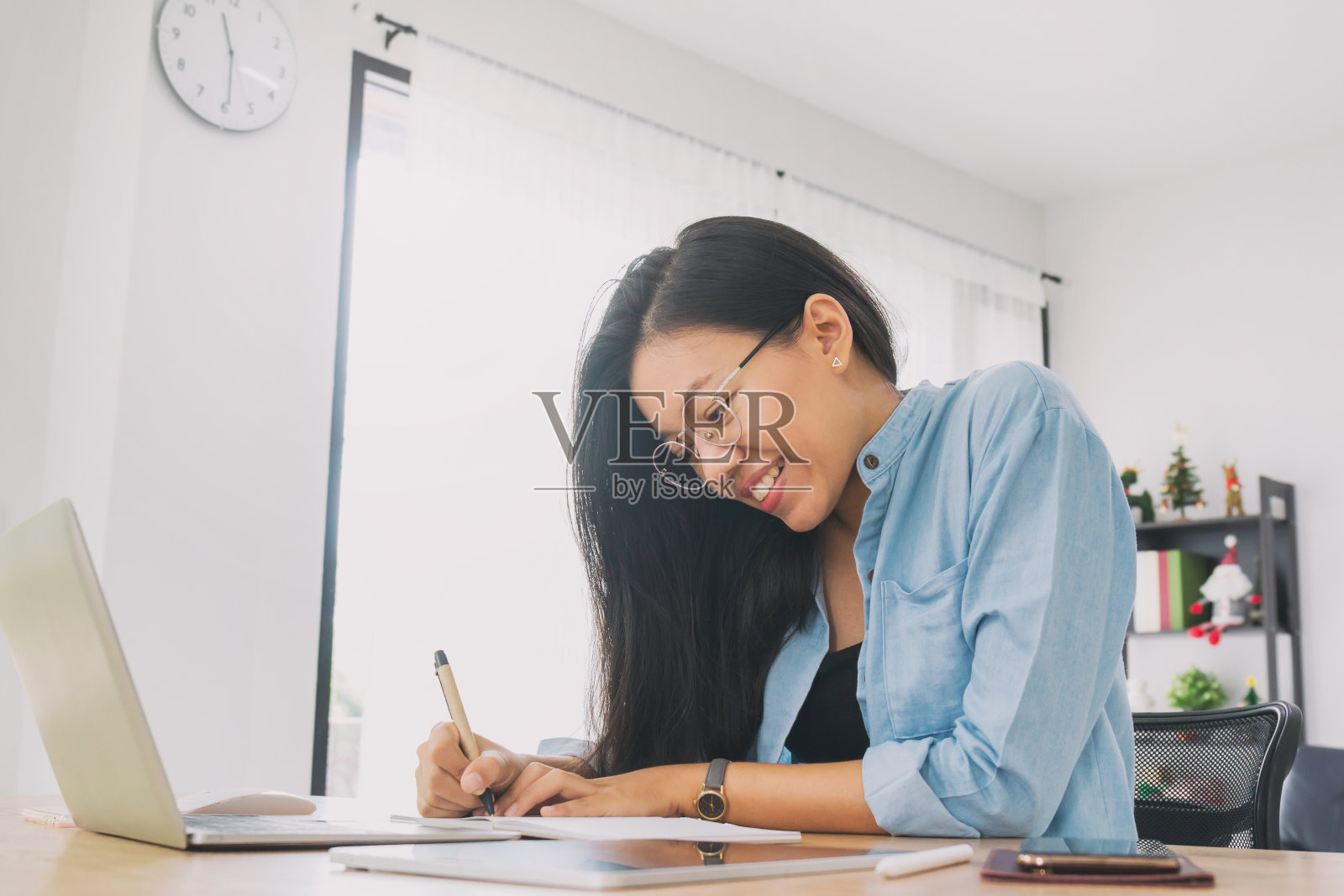 年轻美丽的亚洲女性工作与笔记本电脑，智能手机和平板电脑在家庭办公室装饰圣诞树的背景。女性自由职业者使用手机技术的概念。照片摄影图片