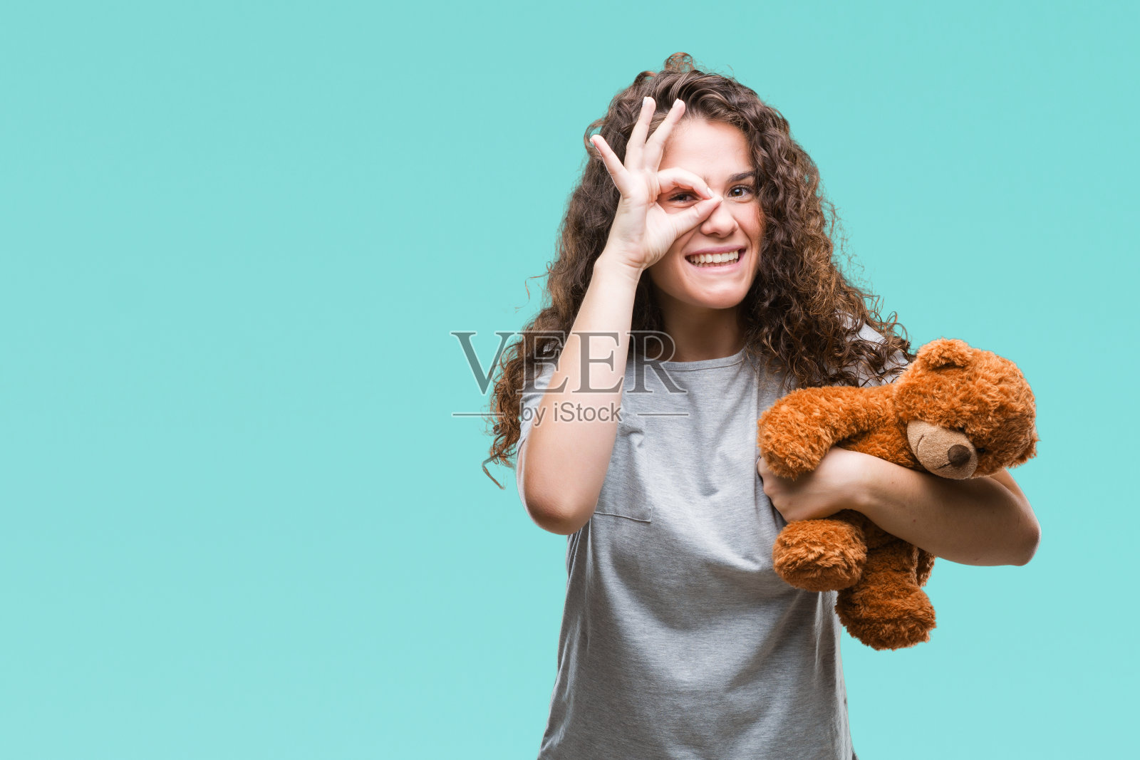 年轻的黑发女孩抱着泰迪熊在孤立的背景与快乐的脸微笑做ok手势与手在眼睛看通过手指照片摄影图片