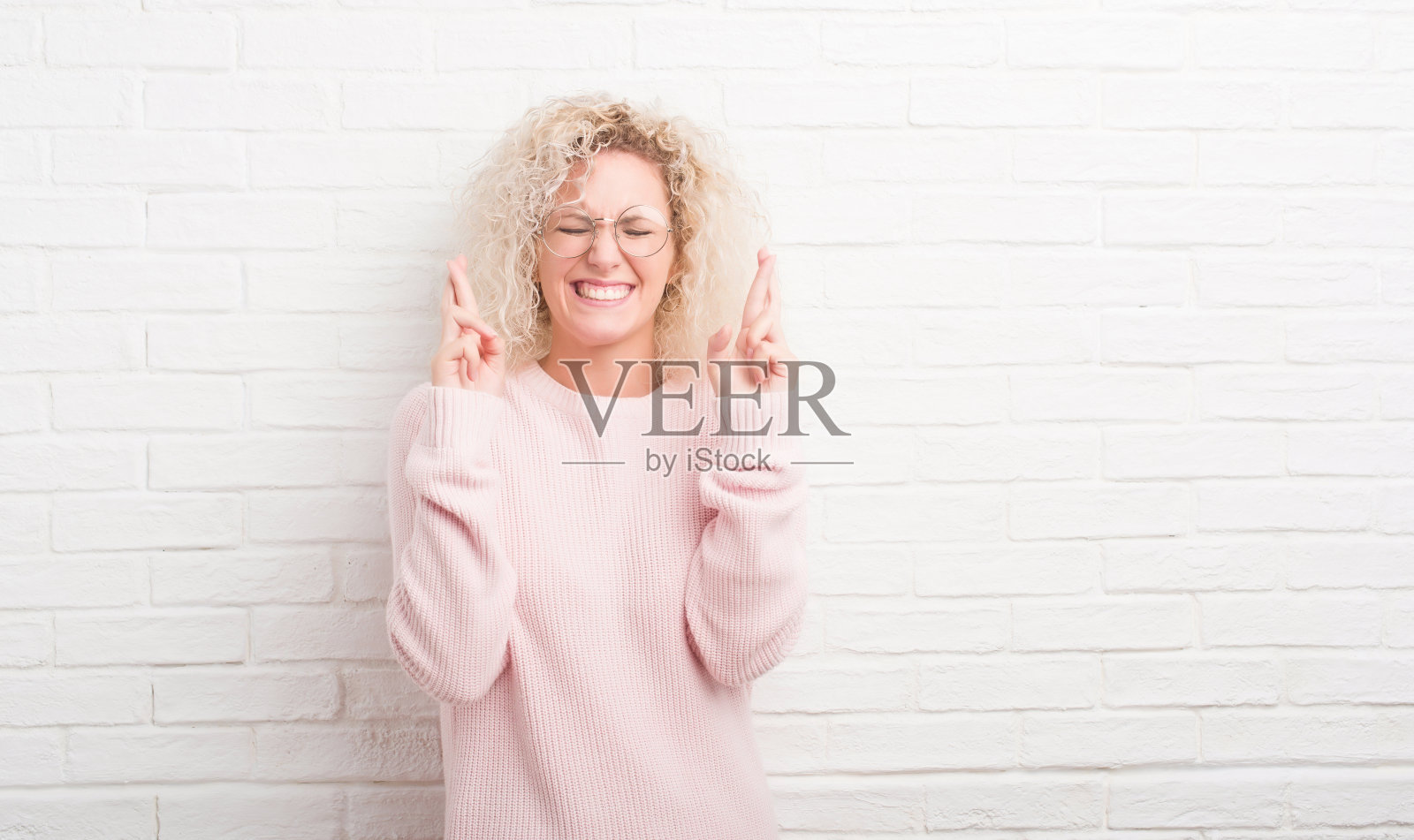 年轻的金发女子，卷着头发，站在白色的砖墙上微笑着，双手交叉着希望，闭着眼睛。运气和迷信的概念。照片摄影图片