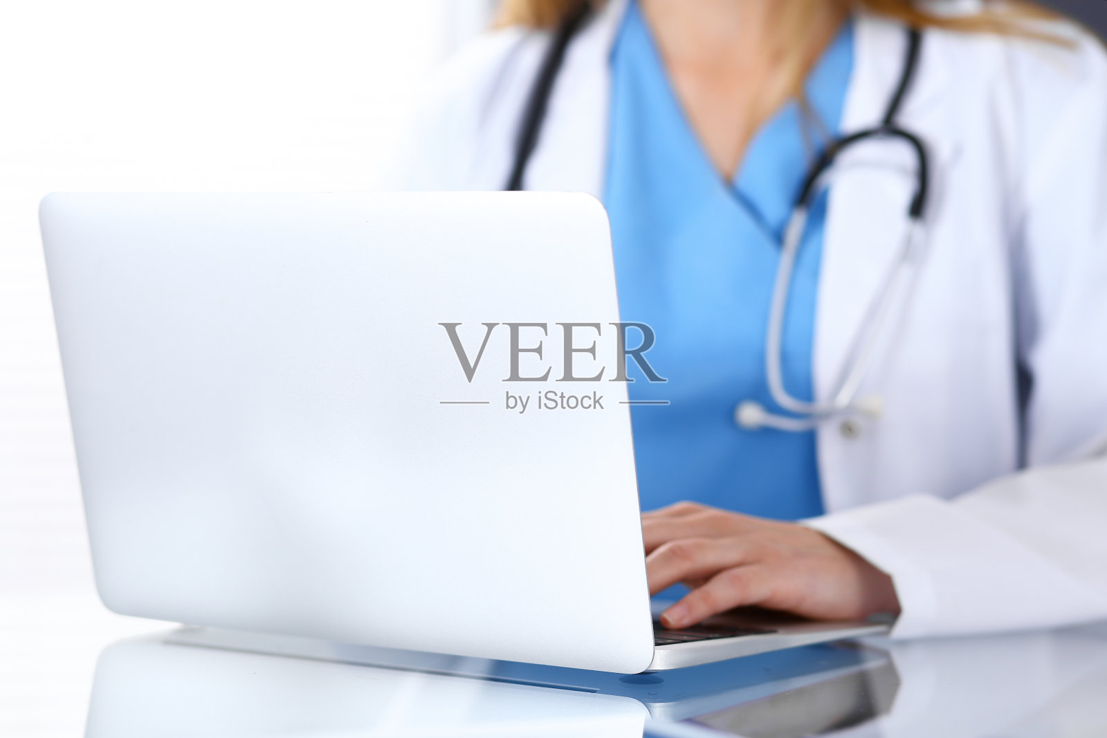 医生坐在医院办公室的玻璃办公桌前用笔记本电脑打字。医生在工作中。医疗保健理念。照片摄影图片