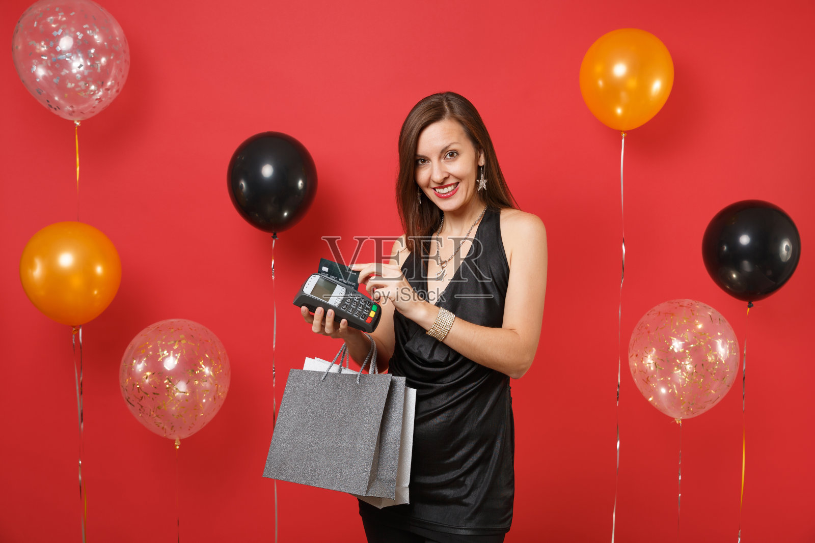 可爱的女孩拿着无线现代银行支付终端，信用卡，包购物后的红色背景气球。新年快乐，生日模型节日派对概念。照片摄影图片
