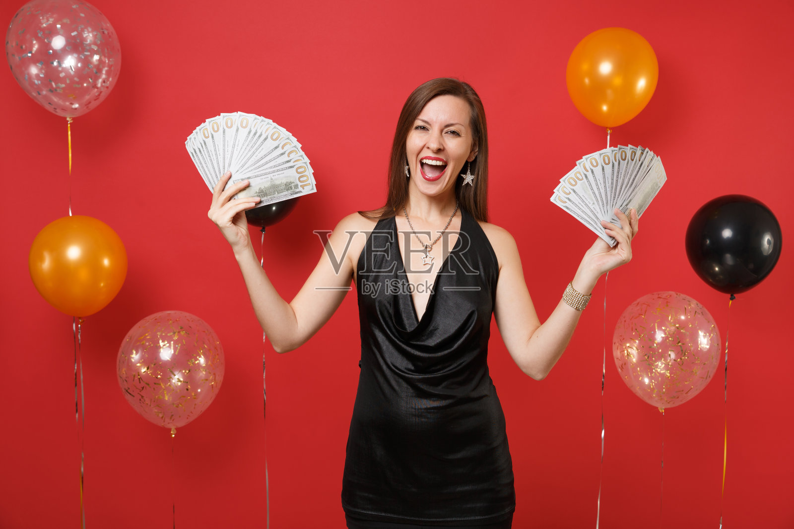 快乐的年轻女子穿着小黑裙，手里拿着一捆现金，放在鲜红色的气球上庆祝。妇女节新年生日模型节日派对概念。照片摄影图片