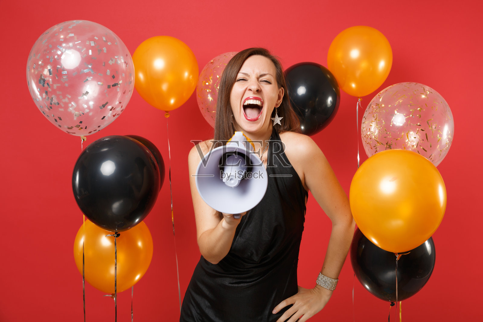 一个穿着小黑裙的疯狂年轻女人在庆祝，用扩音器对着亮红色背景的气球尖叫。情人节，妇女节，新年快乐，生日模型节日派对概念。照片摄影图片